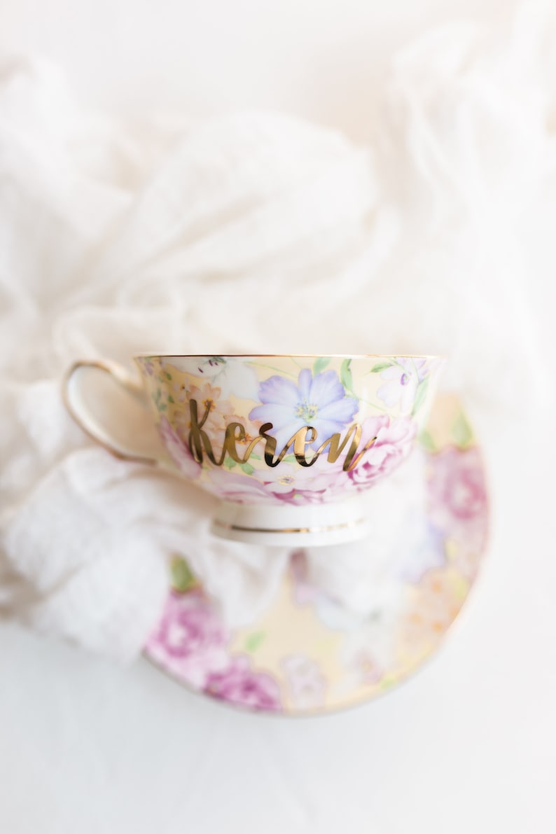 Puede incluir: Una taza de t&eacute; y un platillo con flores y una inscripci&oacute;n dorada que dice "Keren".