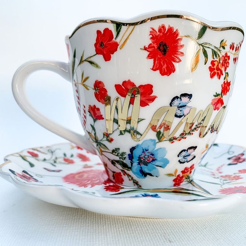 May include: White teacup and saucer with a floral pattern in shades of red, blue, and green. The teacup has a gold rim and the word "Mama" written in gold script.