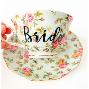 May include: A vintage-style teacup and saucer set with a floral pattern in pink, yellow, and white on a pale green background. The word "Bride" is written in black script on the teacup. The set has gold trim.
