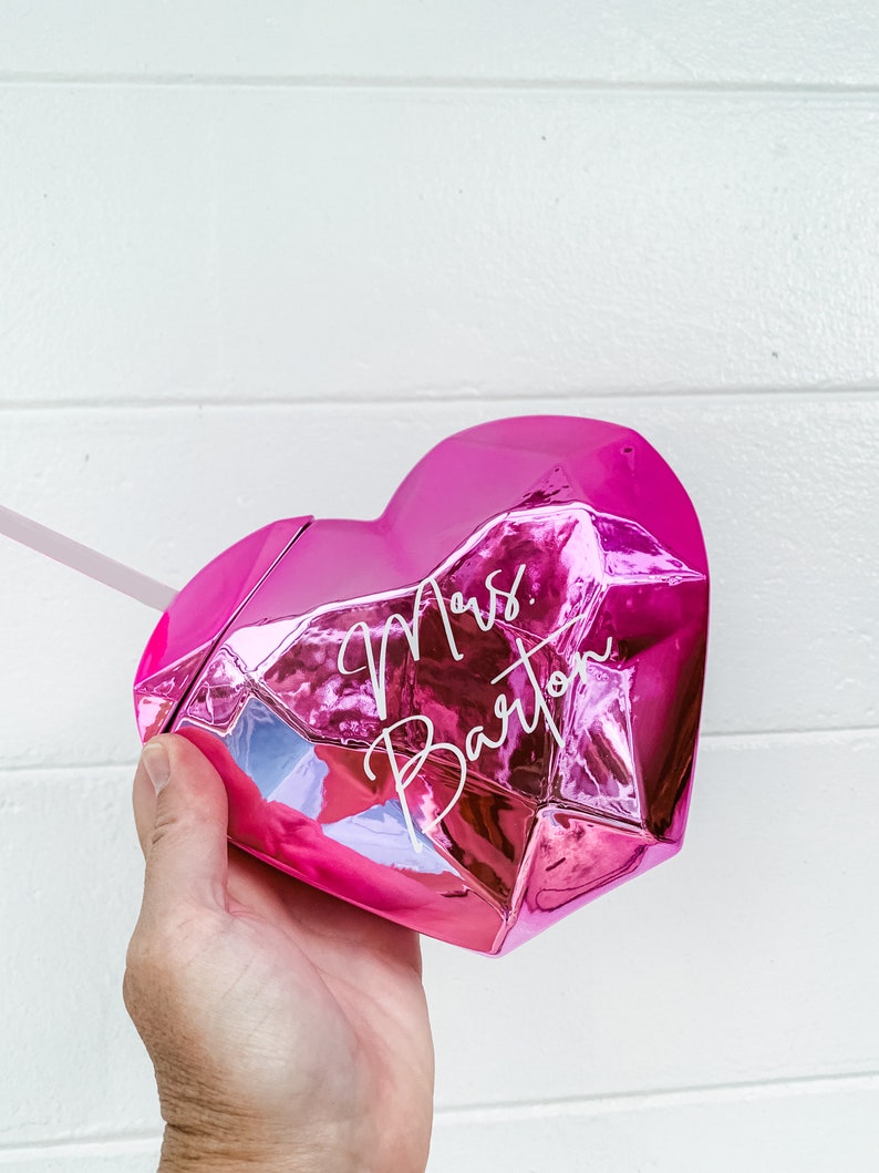 May include: Pink heart-shaped tumbler with a faceted design. The tumbler has the text "Mrs. Barton" written in white on the front.