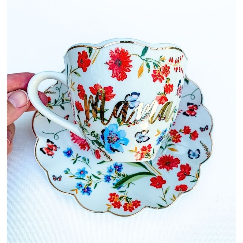 May include: White teacup and saucer with a floral pattern and gold lettering that says "Mama".