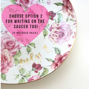 Puede incluir: Un plato de cer&aacute;mica blanco con un patr&oacute;n floral en tonos de rosa y verde. El plato tiene un borde dorado y presenta el texto "Choose option 2 for writing on the saucer too! (5 words max)" en un coraz&oacute;n rosa. La fecha "July 1, 2018" est&aacute; escrita en letras doradas.