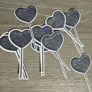 May include: A collection of heart-shaped stickers on sticks. Each sticker has a dark grey heart with the text "SUCKER FOR MORALLY GREY MEN" in a white font. The stickers are on a light brown surface.