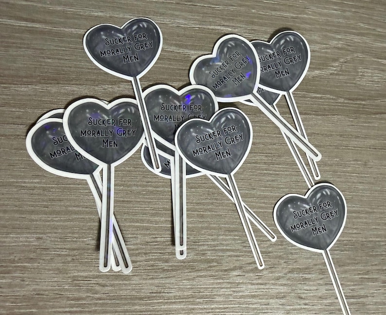 May include: A collection of heart-shaped stickers on sticks. Each sticker has a dark grey heart with the text "SUCKER FOR MORALLY GREY MEN" in a white font. The stickers are on a light brown surface.