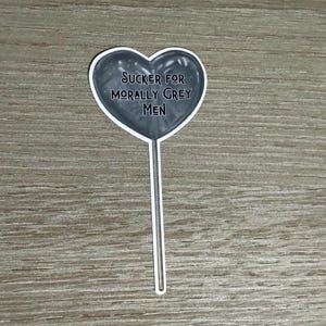 May include: A gray heart-shaped sticker on a white stick. The sticker has the text "Sucker for Morally Grey Men" in a bold, sans-serif font. The background is a light wood grain.