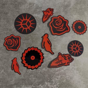 May include: A selection of red and black stickers featuring diverse designs. The stickers depict geometric shapes, saw blades, roses, and stylized figures. The stickers are displayed on a grey surface.