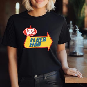May include: Black t-shirt with a graphic design featuring the Vans logo above a yellow arrow with the words "ELDER EMO" in blue text. The design has red and white accents. The shirt is made of a soft material.