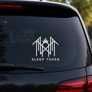 Puede incluir: Un adhesivo de vinilo blanco aplicado en la ventana trasera de un coche oscuro. El diseño presenta un emblema geométrico simétrico con líneas nítidas y angulares y un motivo central de rayos de sol, sobre el texto SLEEP TOKEN en letra sans-serif.