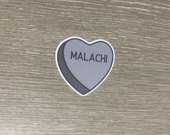 Puede incluir: Una pegatina gris en forma de corazón con el nombre "MALACHI" impreso en negro. La pegatina tiene un borde blanco y un efecto de sombra, sobre un fondo de vetas de madera.