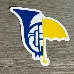 May include: A sticker featuring a blue French horn and a yellow umbrella, both outlined in white. The horn is on the left, and the umbrella is on the right. The sticker is on a wooden surface.
