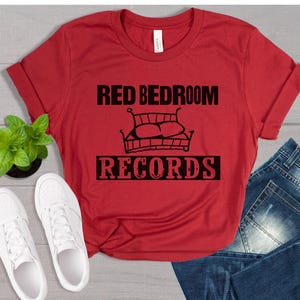 May include: A red t-shirt with the text "RED BEDROOM RECORDS" in black, along with a bed illustration. The shirt is paired with blue jeans and white sneakers, suggesting a casual, everyday style.