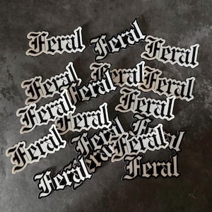 May include: A collection of white and black stickers with the word "Feral" in a gothic font. The stickers are scattered across a dark gray surface, with the word "Feral" in a bold, stylized font.