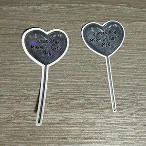 May include: Two heart-shaped stickers with the text "Sucker for Morally Grey Men". The hearts are dark grey with a white border and a stick attached. The stickers are on a wooden surface.