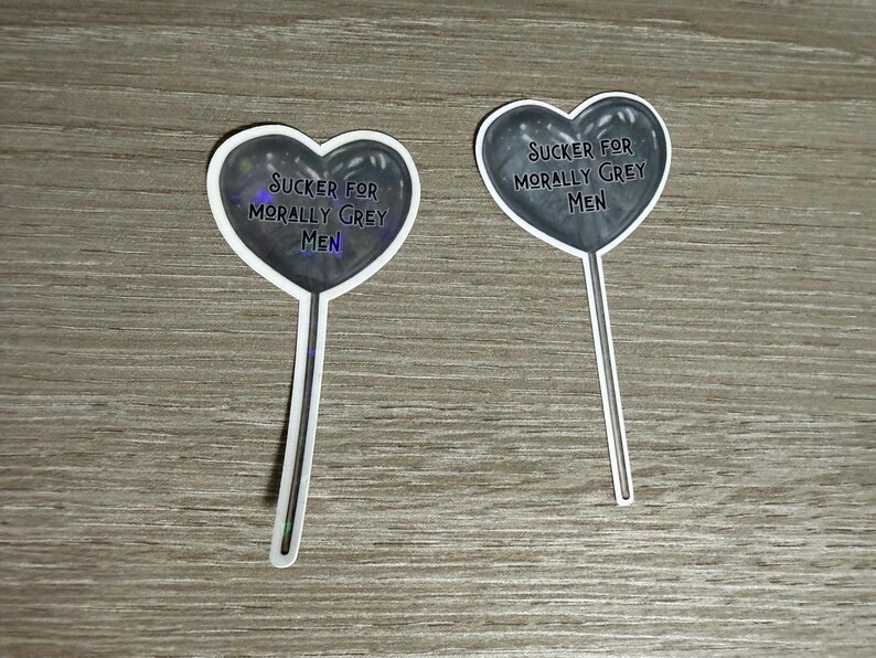 May include: Two heart-shaped stickers with the text "Sucker for Morally Grey Men". The hearts are dark grey with a white border and a stick attached. The stickers are on a wooden surface.