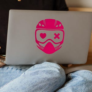 May include: A gray laptop with a bright pink sticker of a helmet with a heart and an X on the visor. The laptop is open and resting on a person's lap, with blue jeans visible.
