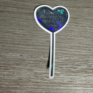 May include: A heart-shaped sticker with the text "SUCKER FOR MORALLY GREY MEN" in a dark font. The heart has a dark, iridescent background with a white border and a white stick.