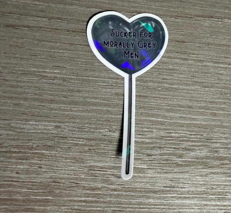 May include: A heart-shaped sticker with the text "SUCKER FOR MORALLY GREY MEN" in a dark font. The heart has a dark, iridescent background with a white border and a white stick.