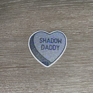 May include: A heart-shaped sticker with the text "SHADOW DADDY" in black capital letters. The sticker has a silver glittery background and a white border. The sticker is on a wooden surface.