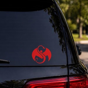 May include: A red vinyl decal applied to the rear window of a black vehicle. The decal features a stylized circular emblem with a central dragon-like figure forming an S-shape, characterized by curved lines and pointed wing-like extensions.