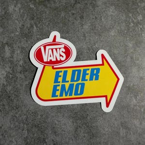 Pode incluir: Um autocolante com uma borda branca e um design de seta amarela. O autocolante apresenta o logótipo Vans num oval vermelho e as palavras "ELDER EMO" em texto azul.