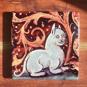 Puede incluir: Azulejo cer&aacute;mico cuadrado con una ilustraci&oacute;n medieval de un gato blanco con ojos amarillos. El gato est&aacute; sobre un fondo rojo oscuro con patrones florales ornamentados de color marr&oacute;n claro. El azulejo tiene un acabado brillante.