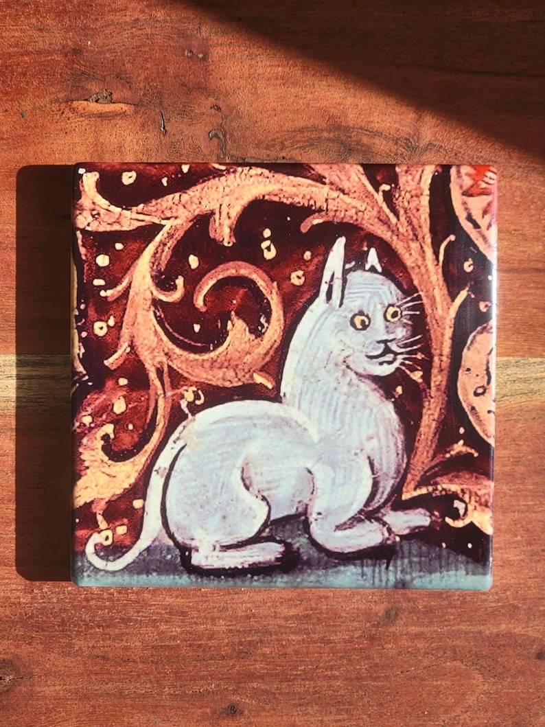 Puede incluir: Azulejo cer&aacute;mico cuadrado con una ilustraci&oacute;n medieval de un gato blanco con ojos amarillos. El gato est&aacute; sobre un fondo rojo oscuro con patrones florales ornamentados de color marr&oacute;n claro. El azulejo tiene un acabado brillante.