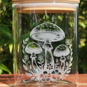Peut inclure: Un bocal en verre transparent avec un couvercle en bois. Le bocal présente un motif gravé de trois champignons entourés de fleurs et de feuilles. Le bocal est conçu pour le stockage et la décoration.