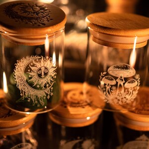 Könnte beinhalten: Klare Glasbehälter mit Bambusdeckeln. Ein Behälter zeigt ein Sonnen- und Mond-Design, während ein anderer eine Pilz-Illustration zeigt. Die Gläser sind wahrscheinlich zur Aufbewahrung gedacht.