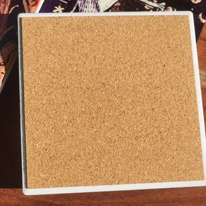 Puede incluir: Posavasos cuadrado con borde blanco y reverso de corcho. El posavasos est&aacute; junto a un posavasos similar con una ilustraci&oacute;n colorida. Los posavasos est&aacute;n sobre una superficie de madera.