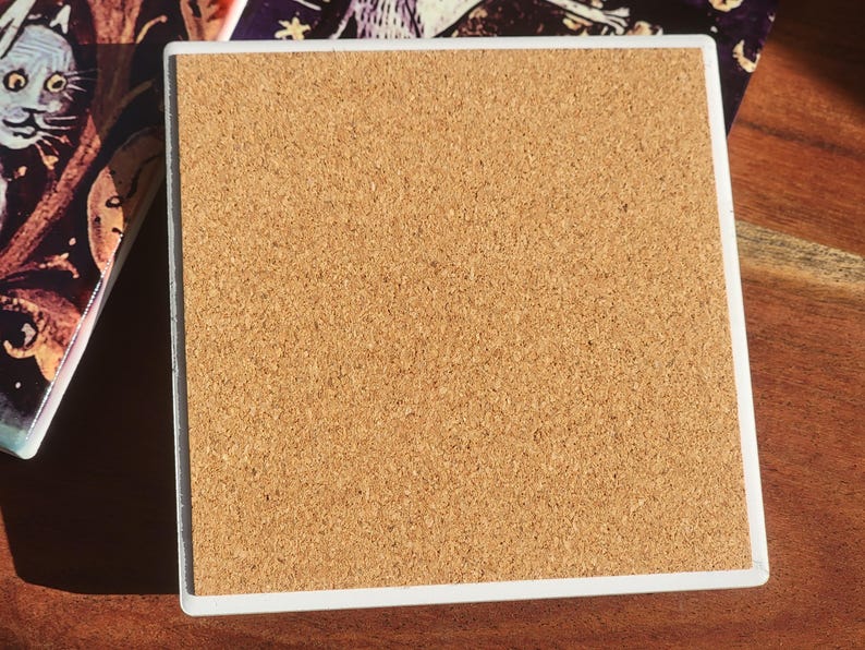Puede incluir: Posavasos cuadrado con borde blanco y reverso de corcho. El posavasos est&aacute; junto a un posavasos similar con una ilustraci&oacute;n colorida. Los posavasos est&aacute;n sobre una superficie de madera.