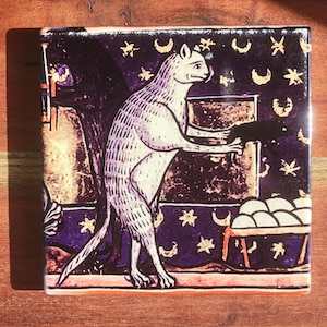 Puede incluir: Azulejo cuadrado de cer&aacute;mica con una ilustraci&oacute;n medieval de un gato. El gato, en tonos blancos y morados, est&aacute; de pie, sosteniendo un objeto oscuro. El fondo es de color p&uacute;rpura oscuro con estrellas y lunas.