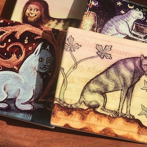 Puede incluir: Un conjunto de posavasos cuadrados con ilustraciones de estilo medieval de gatos y otras figuras. Los posavasos presentan una variedad de colores, incluyendo blanco, marr&oacute;n y negro, con detalles y dise&ntilde;os intrincados. Los posavasos est&aacute;n dispuestos sobre una superficie de madera.