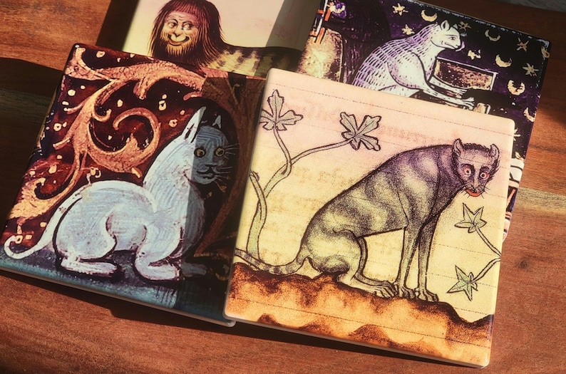 Puede incluir: Un conjunto de posavasos cuadrados con ilustraciones de estilo medieval de gatos y otras figuras. Los posavasos presentan una variedad de colores, incluyendo blanco, marr&oacute;n y negro, con detalles y dise&ntilde;os intrincados. Los posavasos est&aacute;n dispuestos sobre una superficie de madera.