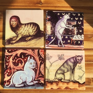 Puede incluir: Cuatro posavasos cuadrados de cer&aacute;mica con ilustraciones de gatos de estilo medieval. Las im&aacute;genes presentan una criatura felina con rostro humano, un gato en una escena de noche estrellada, un gato azul y un animal felino.