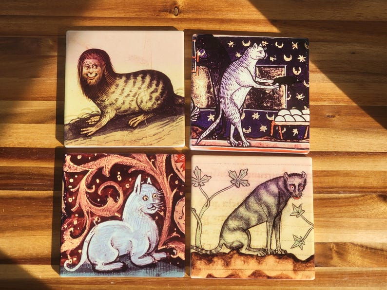 Puede incluir: Cuatro posavasos cuadrados de cer&aacute;mica con ilustraciones de gatos de estilo medieval. Las im&aacute;genes presentan una criatura felina con rostro humano, un gato en una escena de noche estrellada, un gato azul y un animal felino.