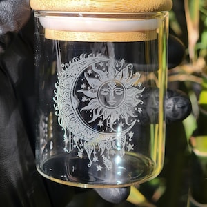 Könnte beinhalten: Ein klares Glasgefäß mit einem Bambusdeckel. Das Gefäß hat ein geätztes Design einer Sonne und eines Mondes mit Sternen. Das Gefäß ist zylindrisch und scheint leer zu sein. Der Deckel ist aus hellbraunem Bambus.