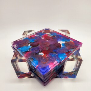 May include: Set of four square coasters with a colorful abstract design in shades of blue, purple, pink, and red. The coasters are made of clear resin and have a glossy finish. The coasters are stacked on top of each other and are held together by a clear acrylic frame.