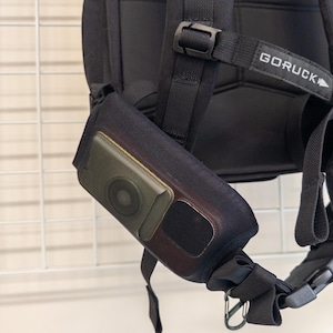 May include: Black backpack with a shoulder strap featuring a small, attached pouch. The pouch has a circular speaker and a rectangular compartment. The backpack has the word "GORUCK" printed on a strap.