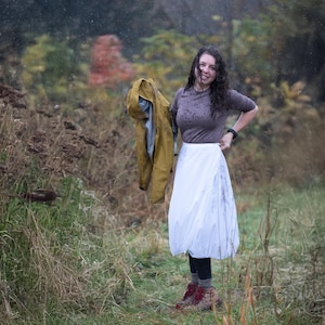 May include: A woman stands in a field, wearing a gray top, white skirt, black leggings, and red hiking boots. She holds a yellow jacket. The background features tall grass, trees, and a rainy sky.