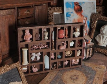 May include: A miniature wooden display case filled with small sculptures and decorative items. The case sits on a wooden surface, with a small rug and other miniature furniture in the background. The items are in various colors.