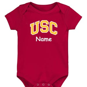 May include: A red baby onesie with short sleeves. The onesie features the text "USC" in yellow block letters with a white outline, and the word "Name" in white, below the letters. The onesie has three snaps at the bottom.