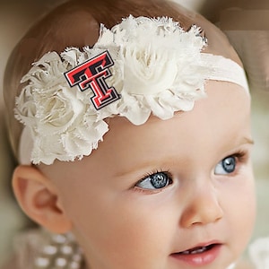 May include: A baby girl wearing a white headband with a white flower and a red and black "T" on it. The baby has blue eyes and is looking to the right.