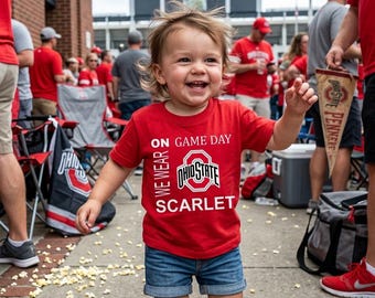 May include: A smiling child wearing a red t-shirt with the text "ON GAME DAY WE WEAR OHIO STATE SCARLET" and blue denim shorts. The child is in a crowd of people, with a stadium in the background.