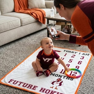 May include: A baby wearing a maroon onesie and matching shoes sits on a white milestone blanket with orange trim. The blanket has the text "FUTURE HOKIE" and a cartoon turkey. A person is taking a photo of the baby.