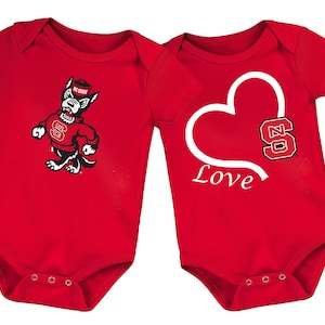 May include: Two red baby onesies. One features a cartoon wolf in a red sweater with the letters "NC State". The other has a white heart with the word "Love" and the letters "NC State" in a red and black design.