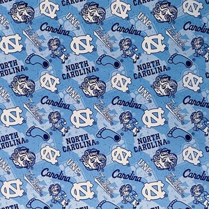 May include: Light blue fabric with a repeating pattern of white logos, text, and illustrations. The design includes the words "UNC", "Carolina", "Tar Heels", and "North Carolina", along with a ram mascot, a running figure, and a footprint.