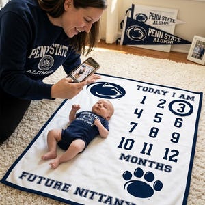 May include: A white baby milestone blanket with navy blue trim featuring the Penn State logo, the text "TODAY I AM" with numbers 1 through 12, the word "MONTHS", and "FUTURE NITTANY LION" at the bottom, accompanied by a navy blue paw print graphic.