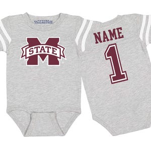 May include: A gray baby bodysuit with white stripes on the sleeves. The front features a maroon and white "STATE" logo. The back displays the word "NAME" above the number "1" in maroon.