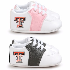 May include: Two pairs of baby sneakers. One pair features white and pink accents, the other white and dark gray. Each shoe has a white base, a colored stripe, and the Texas Tech logo in orange and black.