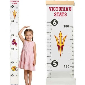 May include: A white hanging growth chart with wooden top and bottom rails, featuring the text VICTORIA'S STATS, a yellow and maroon trident logo, and dual measurement scales in feet and inches up to 6 feet and centimeters up to 180 cm.